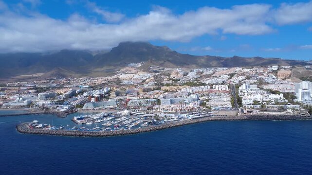 Dron vuela desde el mar hacia Puerto Col&oacute;n y la localidad tur&iacute;stica blanca de Costa Adeje con monta&ntilde;as al fondo, Tenerife, Espa&ntilde;a