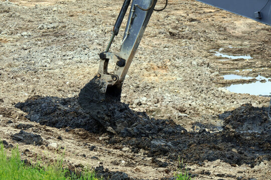 Heavy excavator arm and bucket engaged in earthmoving, extracting dark wet soil from uneven terrain with puddles and sparse grass