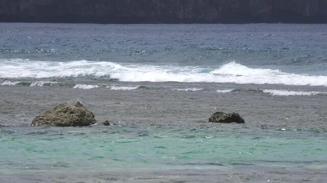 Ocean wavelets rolling over a shallow turquoise reef with exposed volcanic rocks near a dark cliff shoreline.