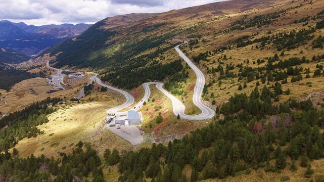 Aerial view of serpentine switchback road winding through golden autumn hillsides in the Pyrenees mountains, with pine forests, valleys and small buildings visible below