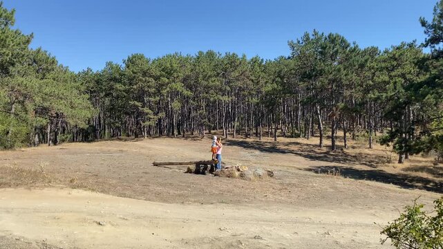 A mother holds her young son as he balances on a fallen log in a large, sunny clearing. They enjoy exploring nature surrounded by a dense pine forest under a clear blue sky.