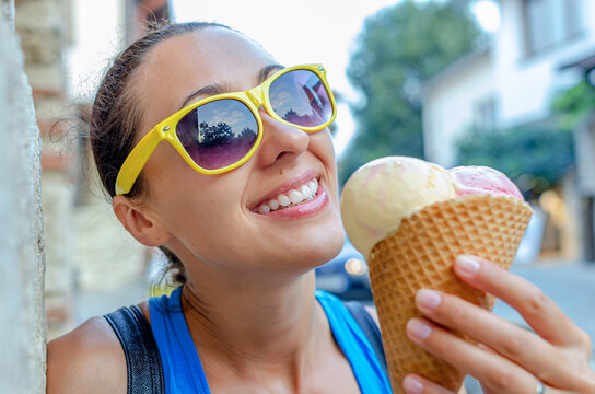 A cheerful young woman in yellow sunglasses holds a cone of ice cream on a sunny day.