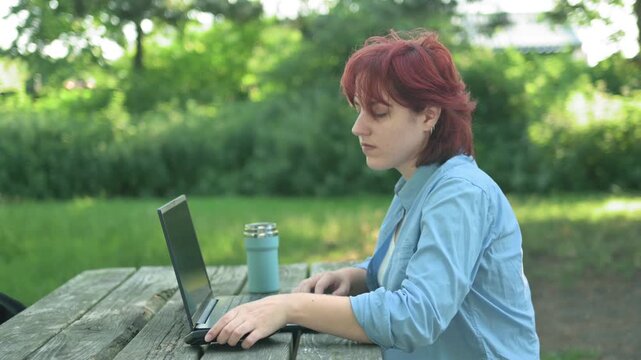 Woman typing text on her laptop at a park table, then stopping to pick up her cell phone and read something on the screen. Concept of receiving an important notification, text messaging, or remote bus