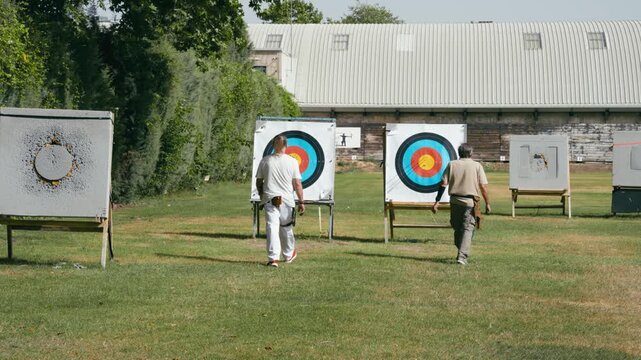 Two archers walking towards their targets on a green field to check the score and retrieve the arrows after a practice session
