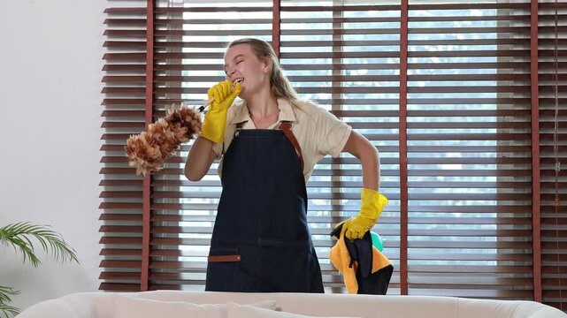 Happy caucasian housekeeper dancing and singing with feather duster while cleaning home furniture and enjoying her domestic work and professional cleaning service job