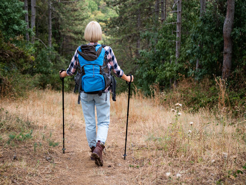 Rear view of happy woman hiker walking along mountain footpath with backpack and hiking pole, exploring nature, wilderness and enjoying outdoor adventure.