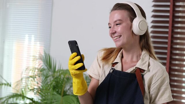 Joyful housekeeper wearing headphones listening to music and singing while using her smartphone, happy janitorial service worker taking a break from cleaning the house