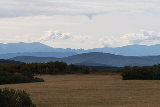 Scenic landscape panorama of harvested fields, autumn forests, and distant blue mountains under cloudy sky   