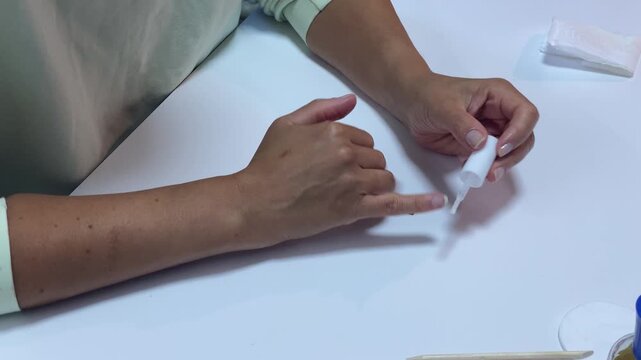 A focused woman carefully applies clear polish to her nail with a small brush. She paints her fingers at a clean white table. This peaceful self care routine gives a feeling of calm.