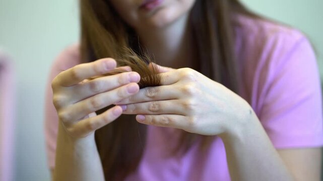 Young woman in a pink shirt holds and closely inspects the damaged split tips of her long brown hair.