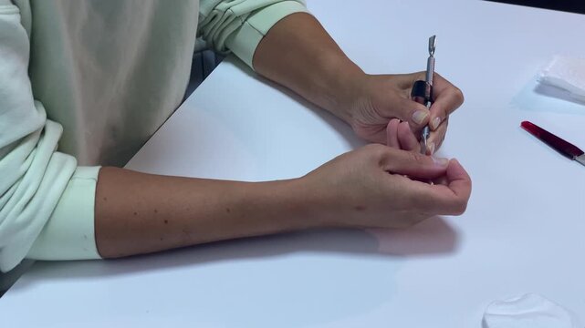 A calm woman gently pushes back her cuticles with a metal tool. She carefully manicures her fingernails at a white desk. This peaceful routine brings a sense of quiet relaxation.