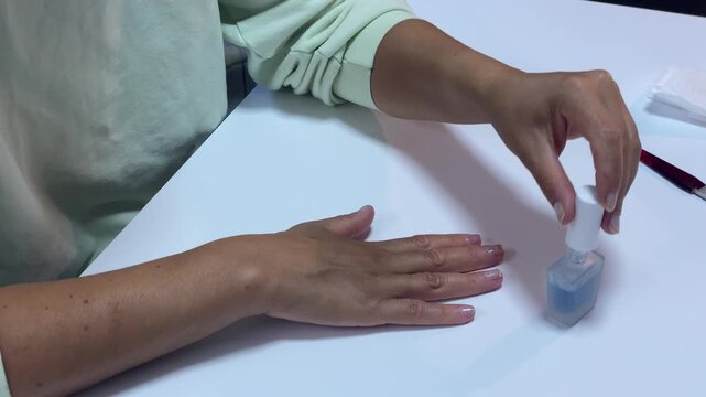 A gentle woman holds a polish brush over a small bottle of blue liquid. She prepares to paint her manicured nails at a clean white desk. This peaceful moment brings a feeling of quiet calm.