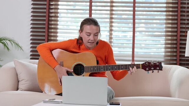 Young smiling woman learning to play acoustic guitar during an online lesson using a laptop, sitting on a sofa in her cozy living room, practicing chords and singing