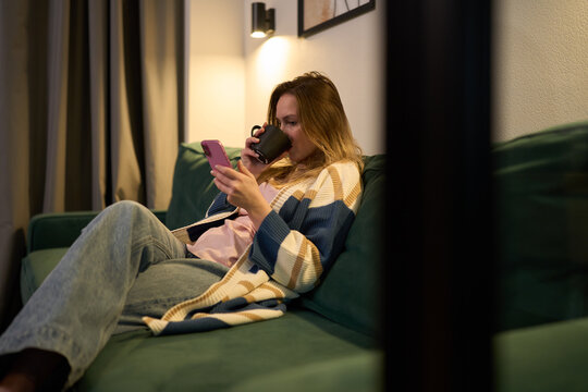 Woman sipping coffee while browsing phone in cozy evening interior
