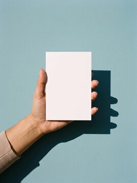 A hand holds a blank white paper flyer against a teal-blue wall with sharp shadows.