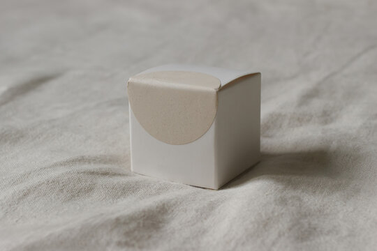 Minimalist White Cubic Gift Box Mockup on Natural Linen Fabric. Elegant Blank Packaging Box with Neutral Blurred Background. Empty beige circular sticker template for logo, text placement. Copy Space
