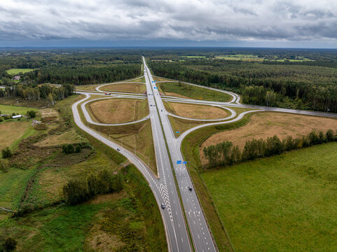 Aerial view of the A7 highway interchange with connecting ramps, divided highway and surrounding forest and agricultural landscape near Ķekava, Latvia.