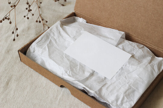 Open Cardboard Gift Box with White Tissue Paper and Blank Greeting Card, Invitation. Business Card Mockup. Minimal Eco Friendly Packaging Template. Neutral blurred Background, dry flowers, Copy Space