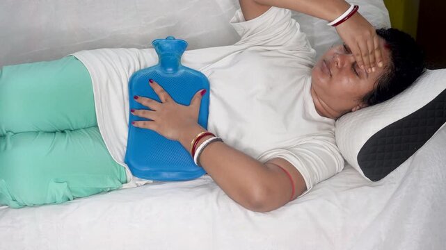 Indian woman suffering from heavy menstrual cramps or periods pain, applying a blue hot water bag on her stomach to relieve the pain and feel better, lying on the living room sofa.