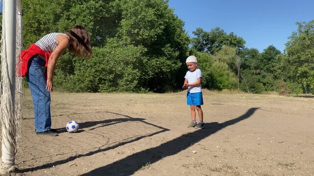 A proud boy gestures excitedly while his mother covers her eyes near a soccer ball. They play on a sunny dirt field surrounded by green trees. The playful game feels full of warmth and cheerful joy.