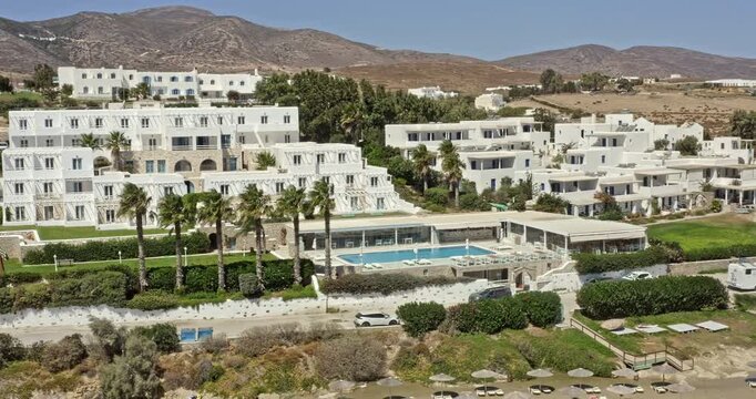 Chrisi Akti Paros Greece Aerial v3 cinematic pan shot, low level fly around hillside beach resorts and summer holiday homes capturing activities of sunbathers and windsurfers - September 2021