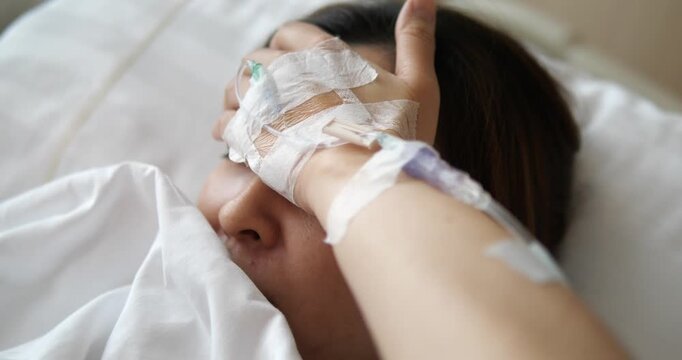 Close up of sick Asian woman covering forehead with IV drip hand on hospital bed