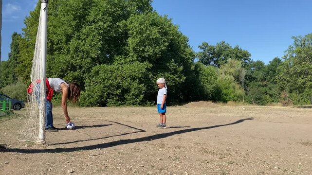 A caring mother sets a soccer ball on the ground for her young boy. The happy child stands watching eagerly on a sunny dirt field surrounded by green trees.