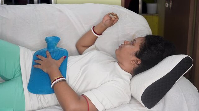 Indian young sick woman checking fever with thermometer during menstrual periods. Using hot water bag to relive cramp or pain, while lying on sofa at home. Fever and illness concept.