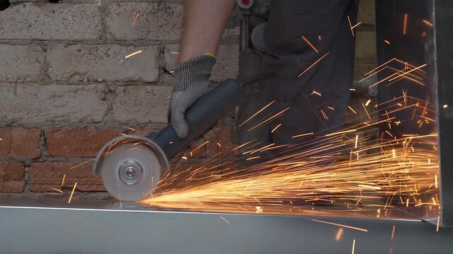 Close-up of a worker cutting a metal sheet with an angle grinder tool in 4K slow motion. Powerful shower of bright glowing sparks flying from the abrasive disc against a brick wall background