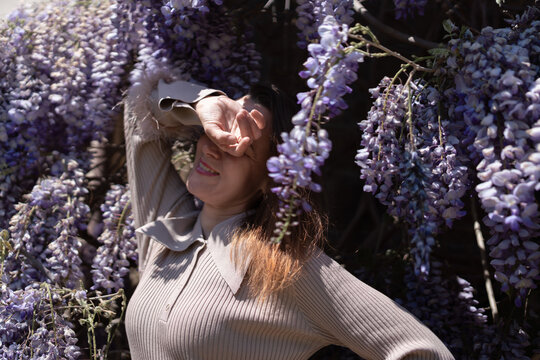 Young woman posing elegantly surrounded by blooming purple wisteria flowers in a sunlit garden during the spring season