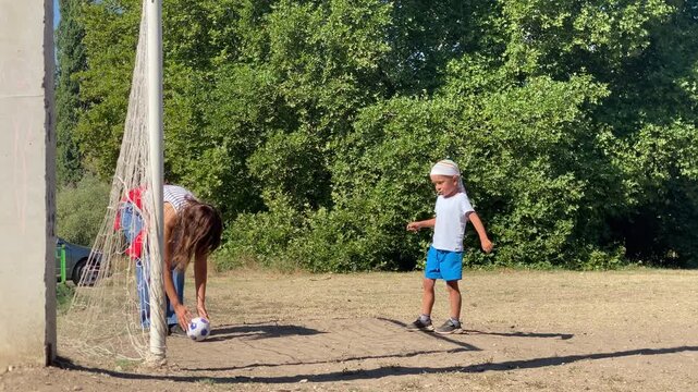 A loving mother bends down to adjust a soccer ball while her excited young son balances playfully on one leg. They share a joyful moment on a sunny dirt field surrounded by green trees.