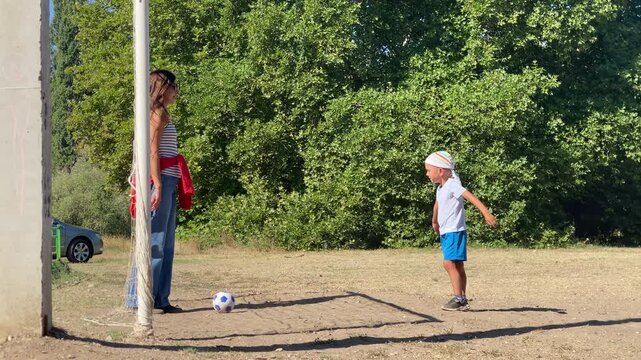 A young boy stands facing a woman who waits near the soccer goal post on a sunny field. They share a peaceful, happy moment playing together outdoors surrounded by green trees.