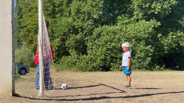 A young boy stands focused as his mother waits near the soccer ball. They share a sweet, quiet moment together on a sunny dirt field with green trees behind them.