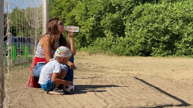 A refreshed mother drinks water while her young boy rests peacefully on a soccer ball. They enjoy a quiet, cozy break together on a sunny dirt field near green trees.