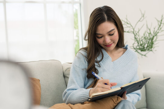 Young Asian woman records daily note in diary book on sofa, relaxed home lifestyle, thoughtful journaling session, weekend planning habit, bright apartment comfort, mindful quiet time indoors.