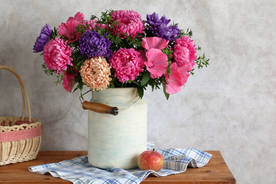 A cottage still life with garden flowers in a canister on the kitchen table. Pink hollyhocks and purple asters. A rustic, cozy composition.