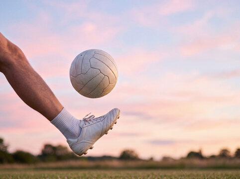 Soccer cleat kicking a white ball high into the air above an open grass field under a soft pink and purple sunset sky. Dynamic sports concept for football, training, energy and motivation.