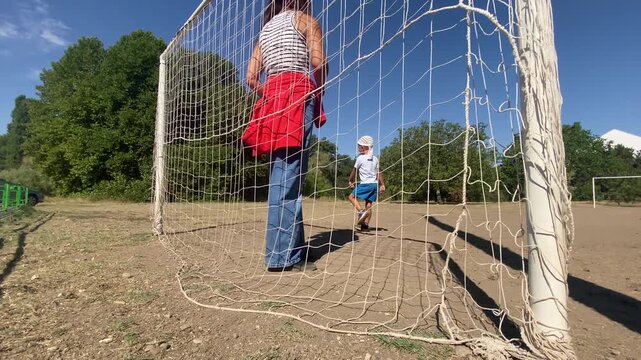 A woman stands inside a soccer goal while a little boy smiles at her. They play together on a sunny dirt pitch. The scene feels happy, warm, and playful.