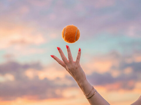 Female hand tossing a small orange ball into the air against a soft pastel sunset sky with pink and teal clouds. A dreamy concept for play, freedom, balance and summer.
