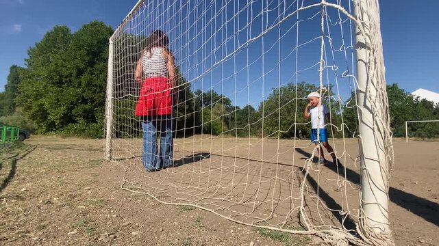 A woman holds a soccer ball inside a goal net while a toddler walks happily nearby. They play a fun football game on a sunny dirt pitch. The warm scene feels cheerful and full of joy.