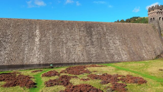 Aerial video capturing the monumental gritstone masonry wall and neo-Gothic valve towers of Derwent Dam towering over the stilling basin and grassy valley slopes in Derbyshire, UK. 