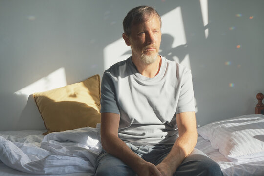 Sad and emotionally exhausted Man sitting on bed, looking away, reflecting during sunlit morning