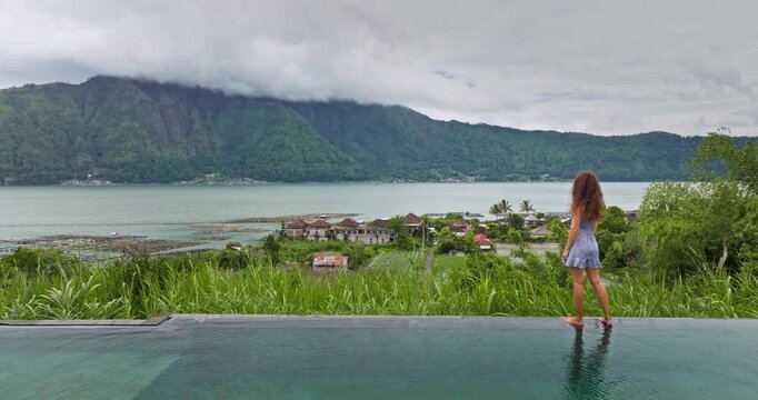 Woman standing at infinity pool edge overlooking Lake Batur and lush green landscape of Bali rural village, enjoying the serene volcanic caldera view on a cloudy day. Travel and explore Indonesia