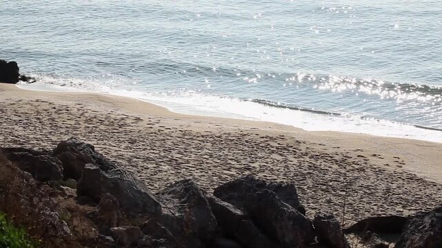 Pan shot of a beach showing ocean waves, sand, and rocks on a sunny day. The sun reflects off the water creating glittering highlights, while the sand is interrupted by large rocks