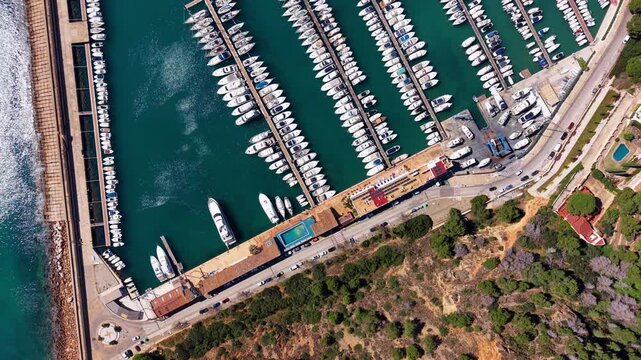 Top-down view of a large Mediterranean marina filled with rows of white boats and yachts moored at parallel docks, surrounded by turquoise water and coastal road