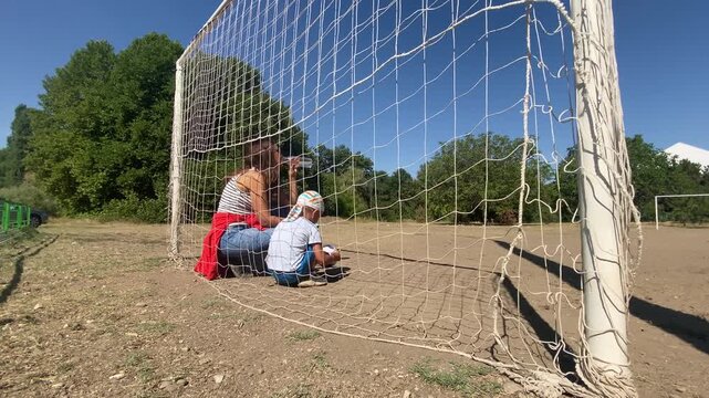 A woman drinks water while crouching inside a soccer goal alongside a young boy taking a rest break. They enjoy a peaceful moment together on a sunny outdoor field filled with loving warmth.