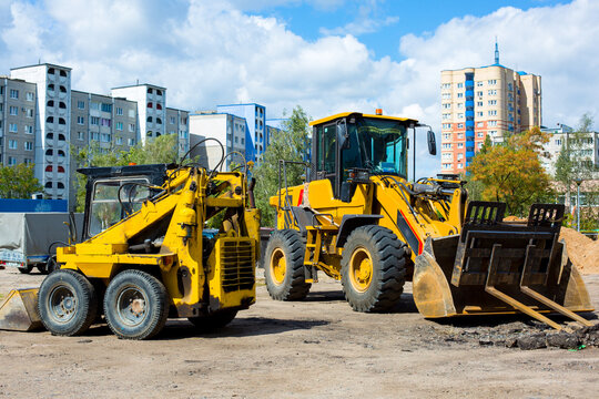 Construction site featuring yellow heavy machinery including a skid steer loader and a wheel loader, with residential buildings visible in the background under a clear blue sky