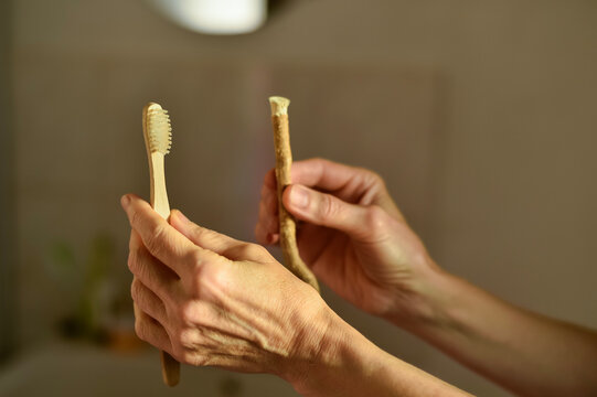 Hands hold a bamboo toothbrush and a natural miswak stick indoors in Paris, France. The comparison shows alternative and traditional dental care tools in a warm bathroom setting.