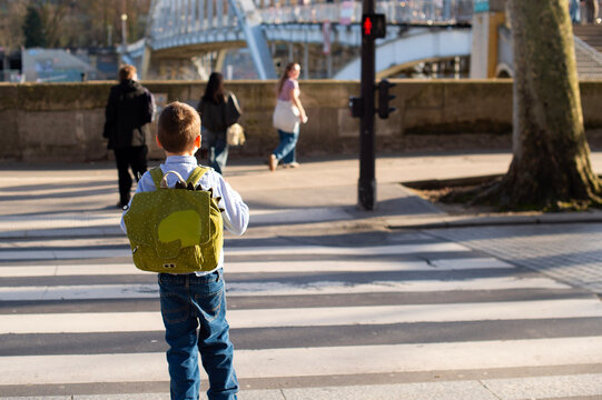 A young boy wearing a green backpack stands at a pedestrian crosswalk in Paris, France. A red pedestrian traffic signal and an arched bridge appear in the background.