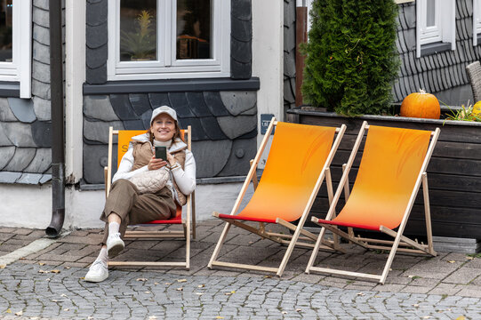 Smiling woman relaxes in deckchair outside slate house in Goslar, enjoying autumn travel mood. Harz tourism, casual fashion and historic streets create cozy lifestyle scene in Germany today